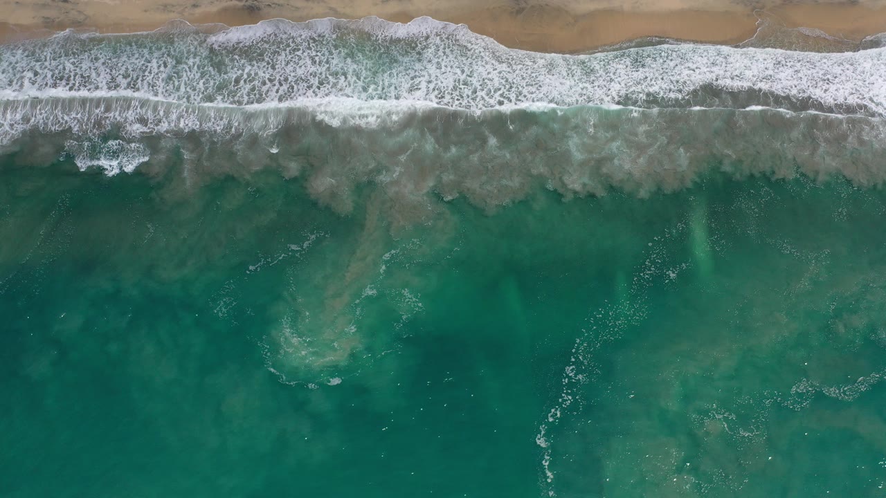 Overhead view of Los Cabos surf with white foam along a narrow sandy beach and green water filling the frame, no visible people.