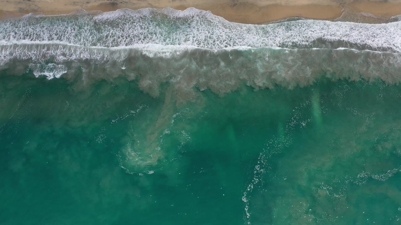A white band of surf advances across turquoise-green water below a sandy Los Cabos beach, with darker underwater patches and no visible people.