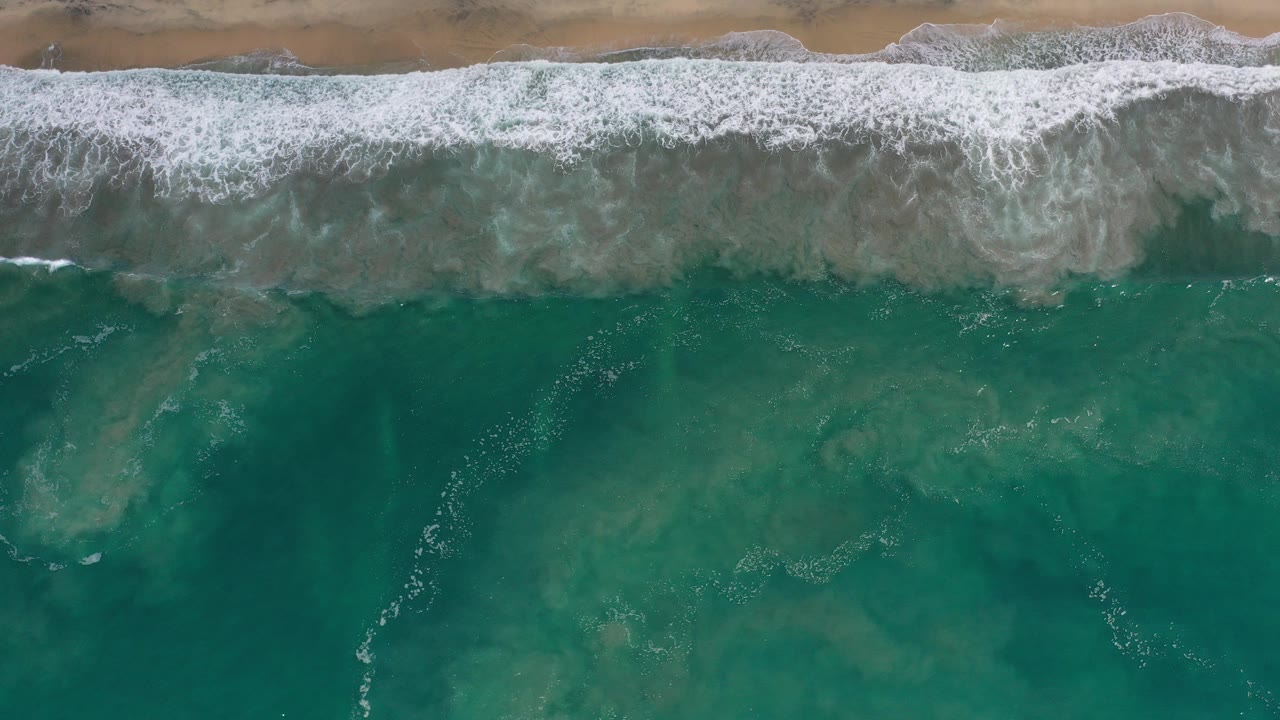 Overhead view in Los Cabos, Mexico showing a broad white surf band breaking along tan sand, blue-green ocean below, darker underwater patches, and no visible people.