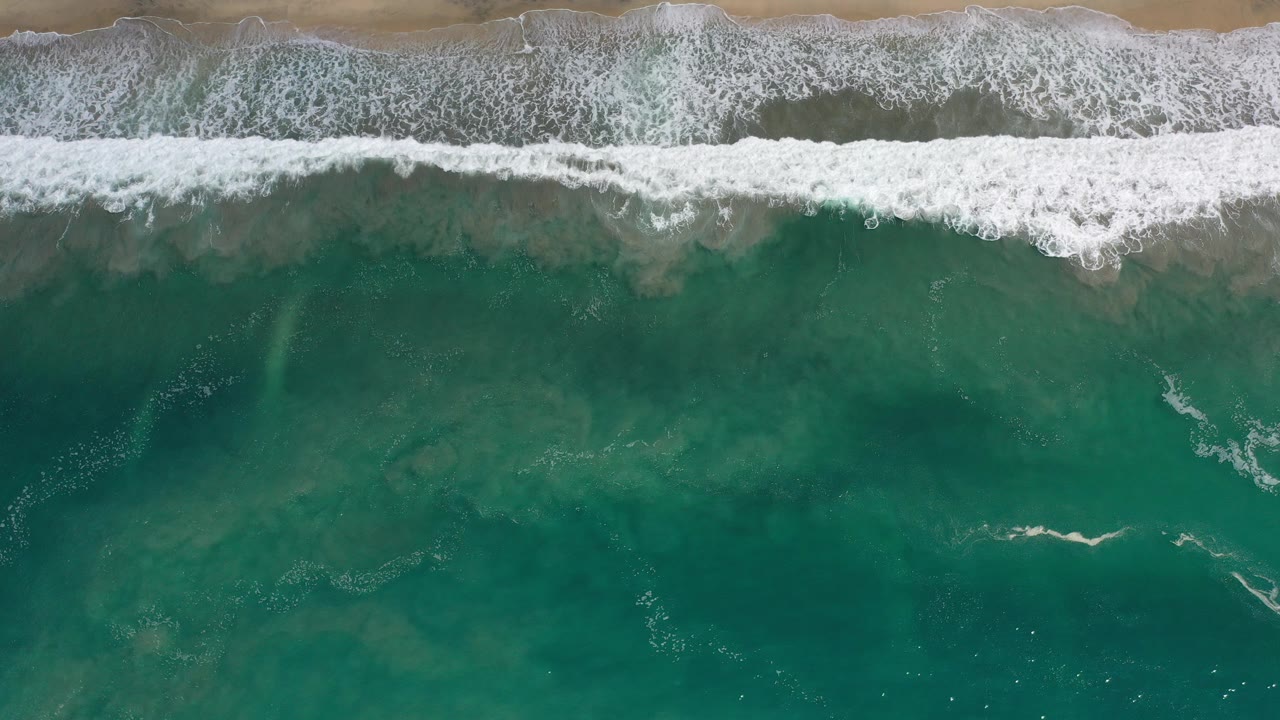 Top-down view of teal water and white surf reaching tan sand in Los Cabos, Mexico, with no visible people.