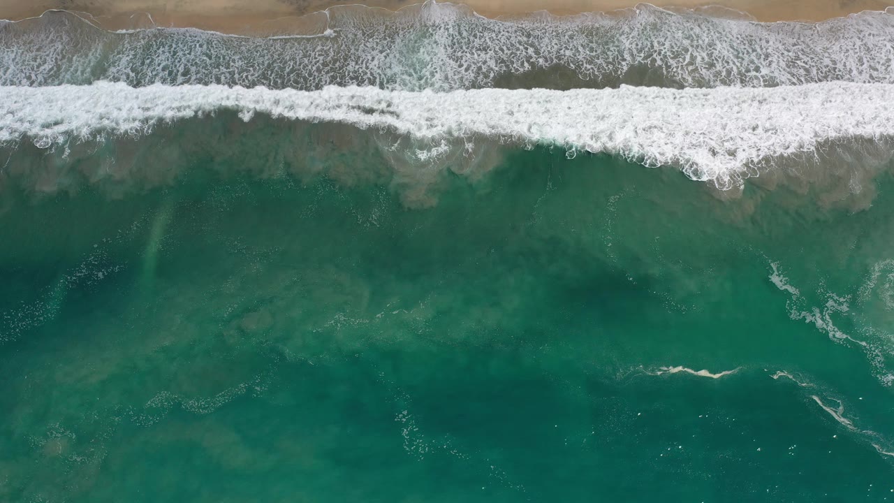 Aerial shoreline with white foam and darker teal water below.
