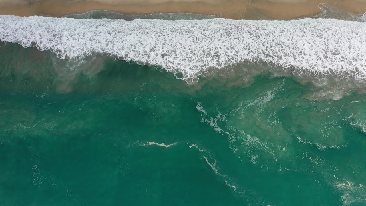 Overhead view in Los Cabos, Mexico, with white surf across the top edge, green ocean below, and no visible people.