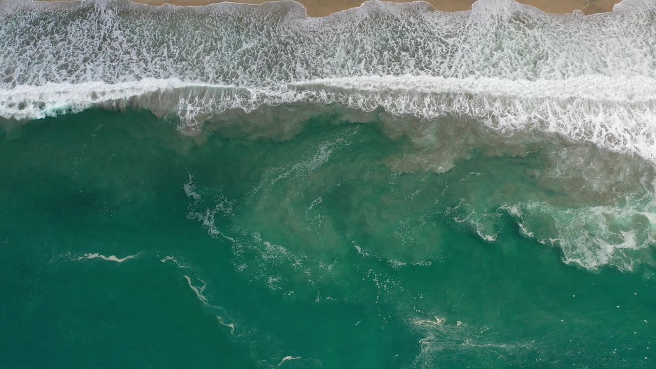 Top-down view in Los Cabos, Mexico, showing white surf breaking across teal-green water with a thin strip of tan sand along the top edge and no visible people.
