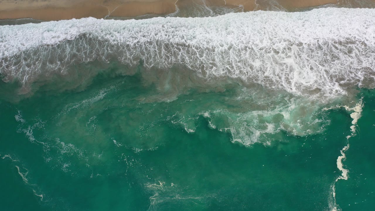 Overhead green ocean surf breaking beside tan sand.