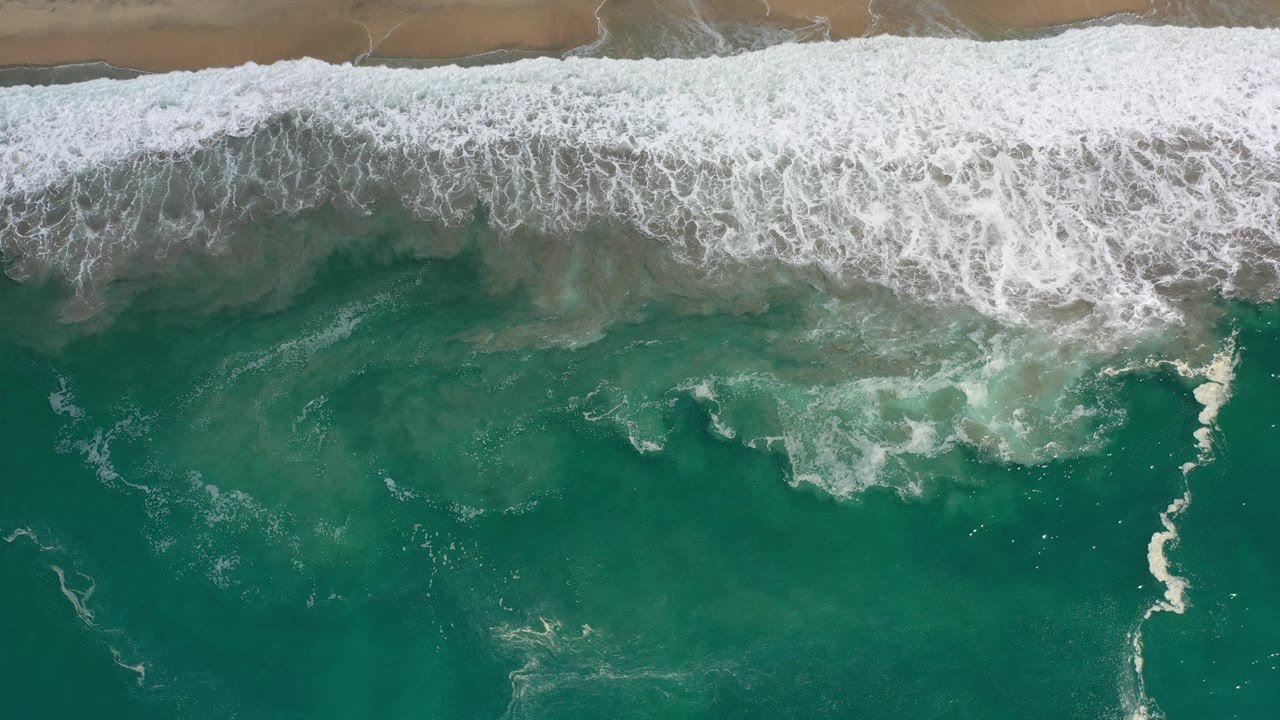 Aerial frame of white foam spreading over green ocean water below a thin tan beach line in Los Cabos, Mexico, with no visible people.