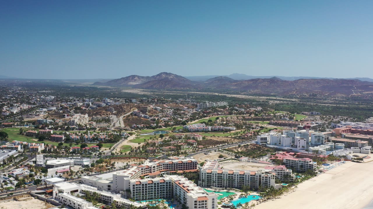 Opening aerial frame over Los Cabos, Mexico, with resort buildings and turquoise pools in the foreground, city roads and greenery beyond, brown hills on the horizon, and no visible people.
