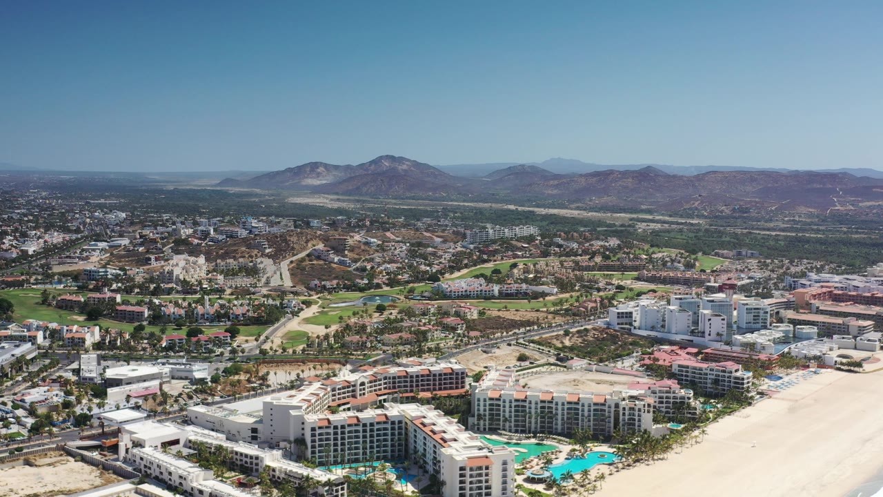 Drone view of beachfront resort buildings, city roads, green areas, and mountains.