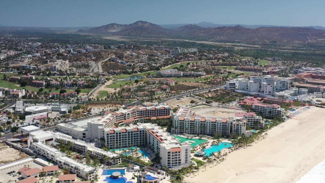 Aerial frame in Los Cabos, Mexico with the beachfront apartment complex centered lower in the view, pale beach to the right, roads and green patches inland, and no visible people.