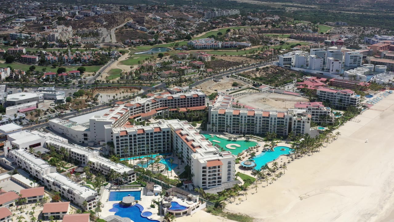 Opening aerial frame over Los Cabos, Mexico, showing a large beachside resort complex, turquoise pools, pale sand to the right, inland roads and buildings beyond, and no visible people.