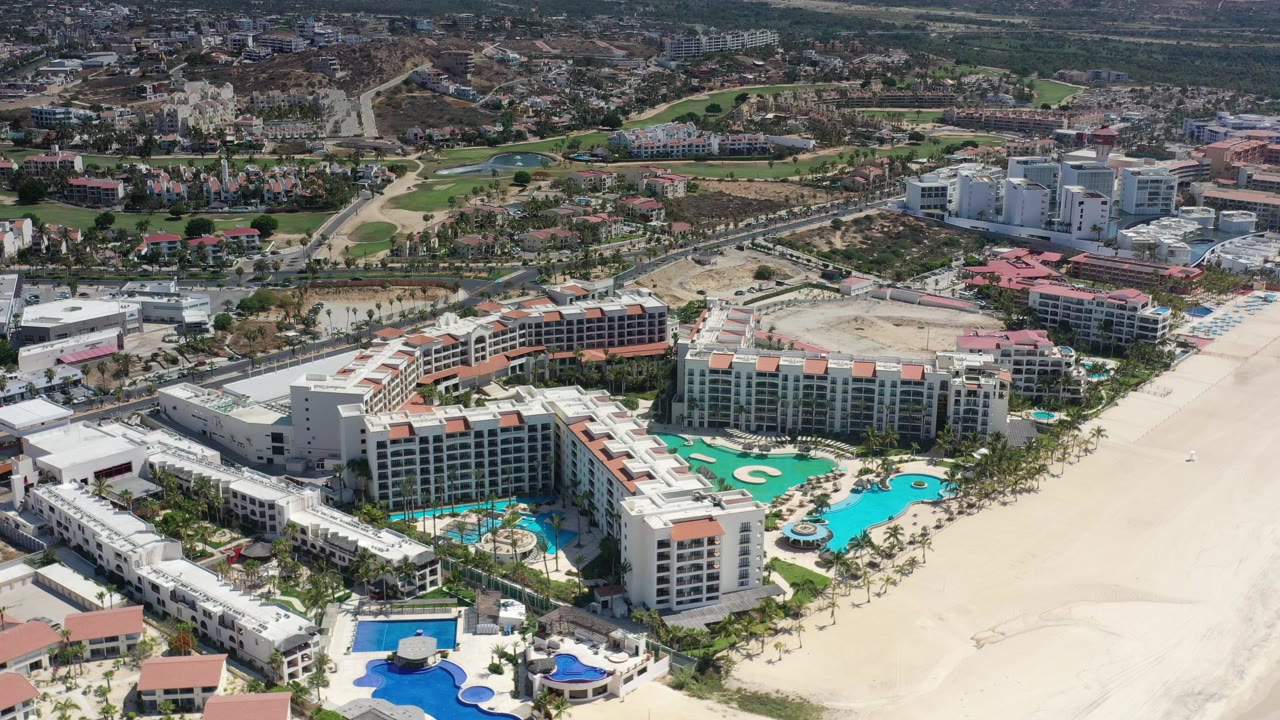 A slightly later aerial view in Los Cabos, Mexico, keeps the resort buildings and bright pool courtyards centered, with the broad sandy beach on the right and no visible people.