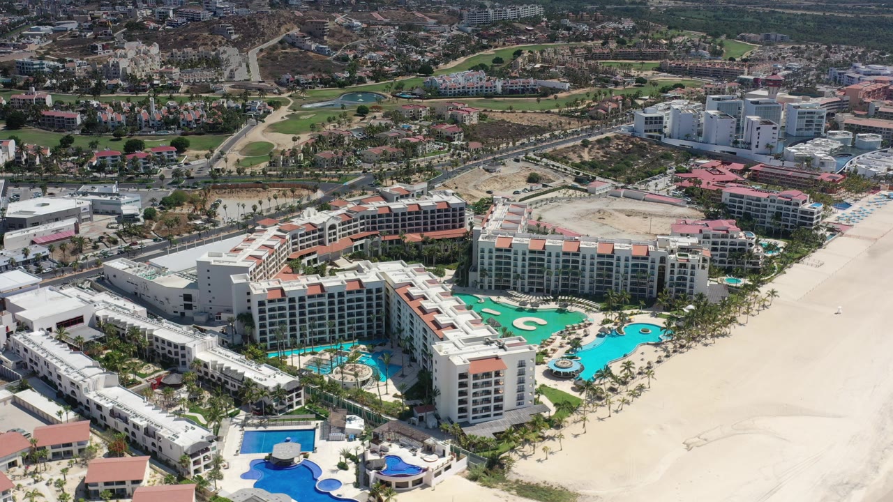 High aerial angle on a coastal hotel complex with pools, beach, roads, and inland development.