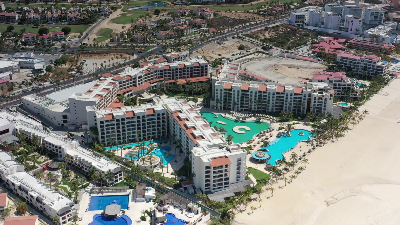 High aerial view in Los Cabos showing a white resort complex with turquoise pools, pale sand, palm rows, and surrounding roads, with no visible people.