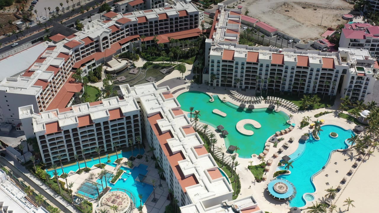 High aerial view in Los Cabos, Mexico showing a broad turquoise pool, adjacent blue pool section, white hotel wings, palms, and no visible people.