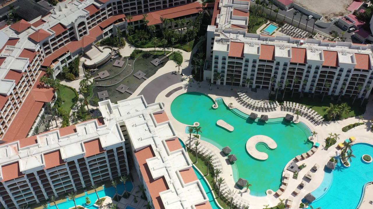 High aerial view over a Los Cabos hotel complex with white buildings, red roof panels, a turquoise pool, lounge chairs, palms, and no visible people.