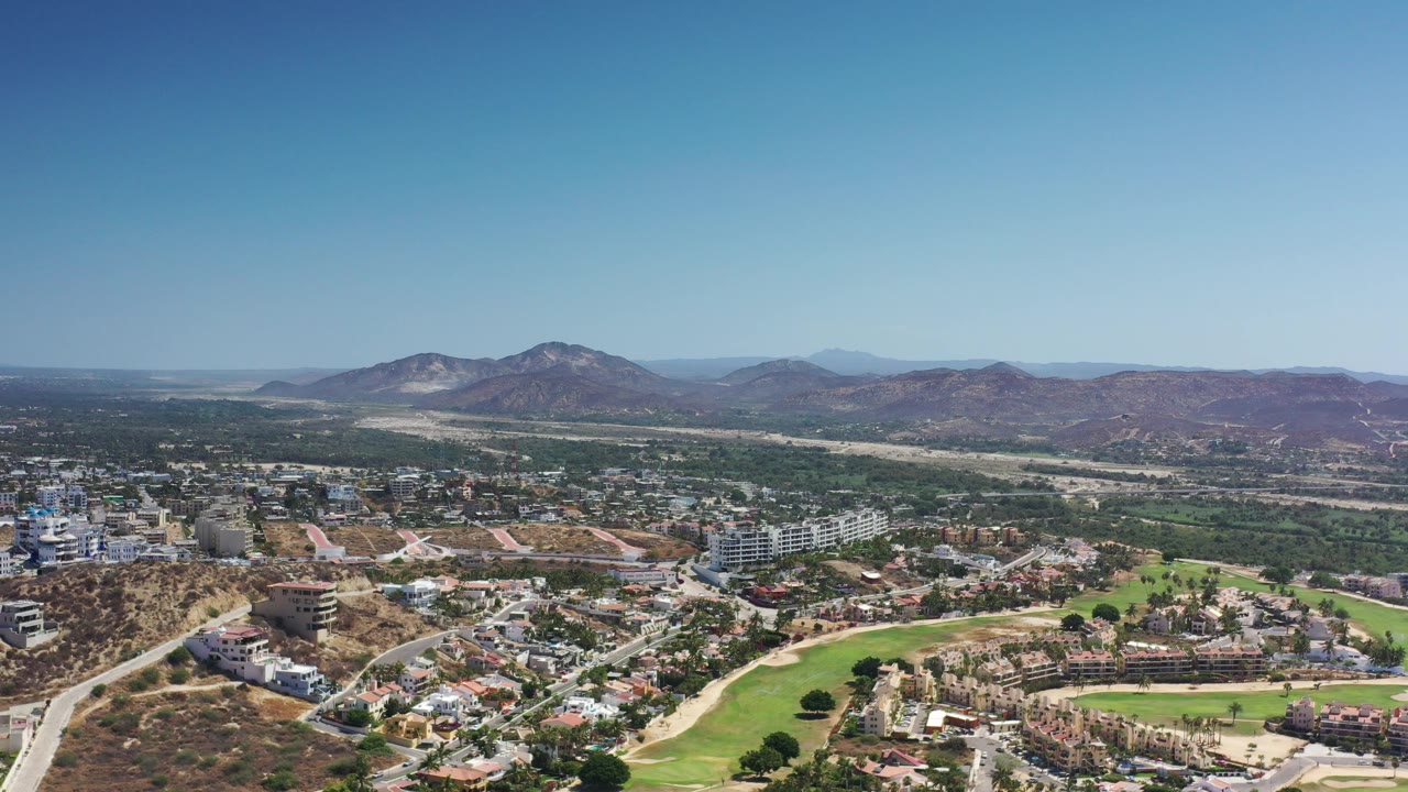 Opening frame of Los Cabos, Mexico, showing a wide aerial view over residential buildings, a green golf course, dry hills, and distant mountains with no visible people.