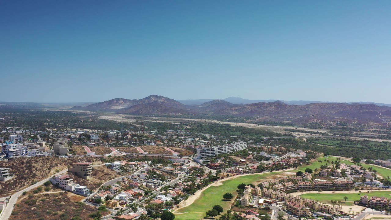 Drone-style view across Los Cabos buildings, fairways, roads, and arid mountains.
