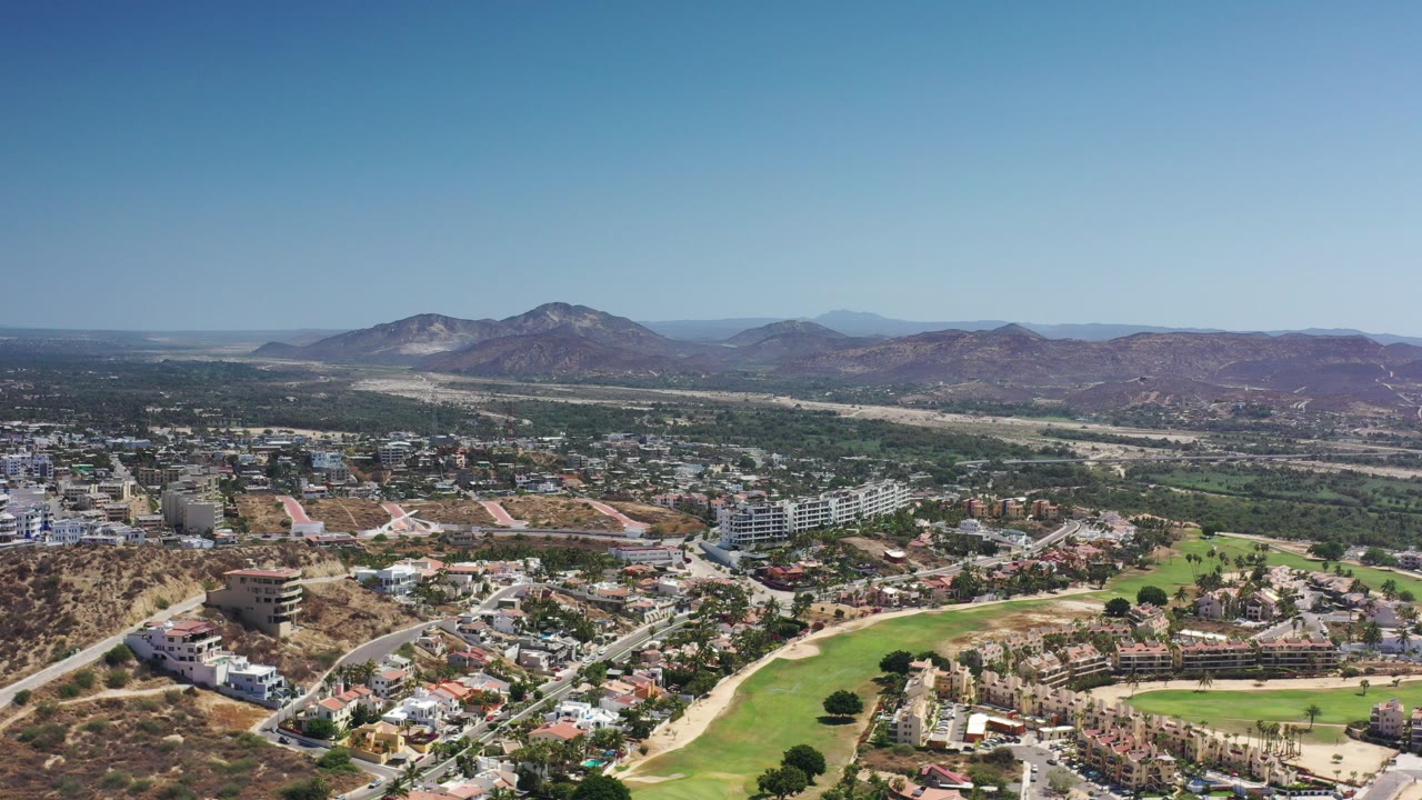 Aerial view of town, golf fairway, dry hills, and distant mountains under blue sky.