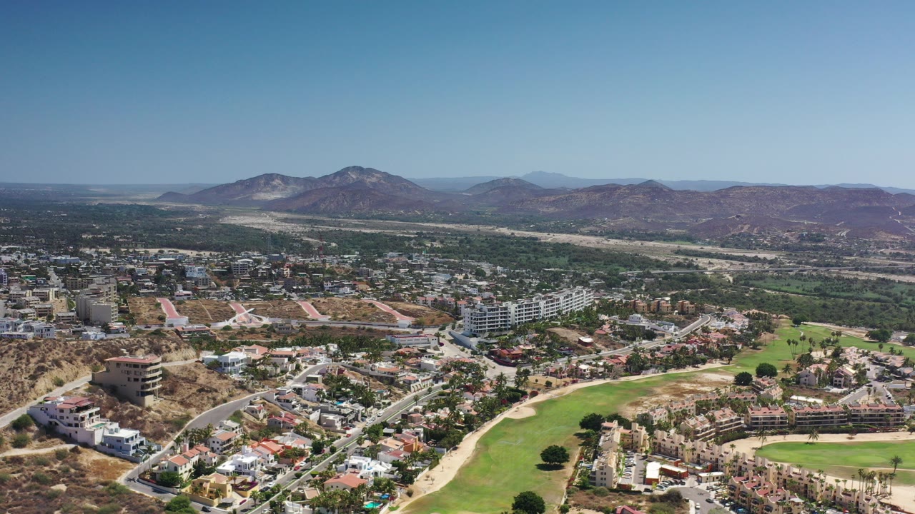 Opening aerial frame over Los Cabos, Mexico, with hillside homes, a bright green fairway, winding roads, dry ridges, distant mountains, and no visible people.