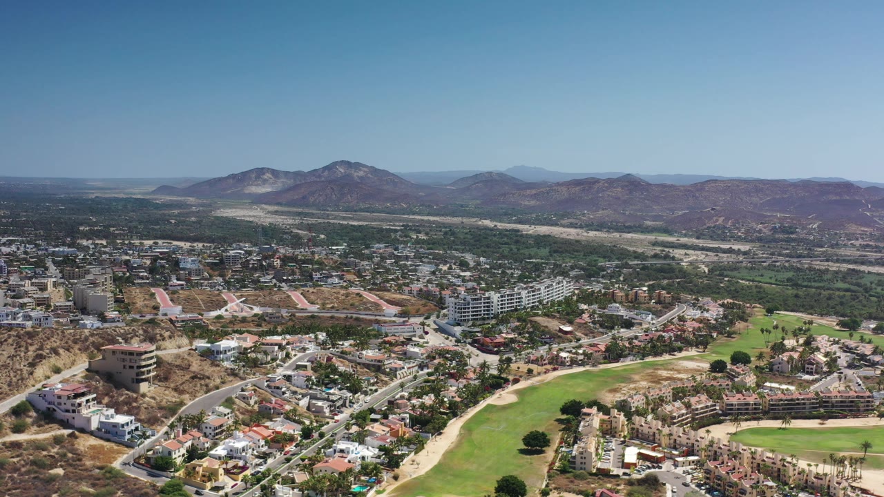 A second aerial view of Los Cabos, Mexico, shows suburban rooftops, curving roads, green golf-course grass, tan hillside terrain, distant mountains, and no visible people.