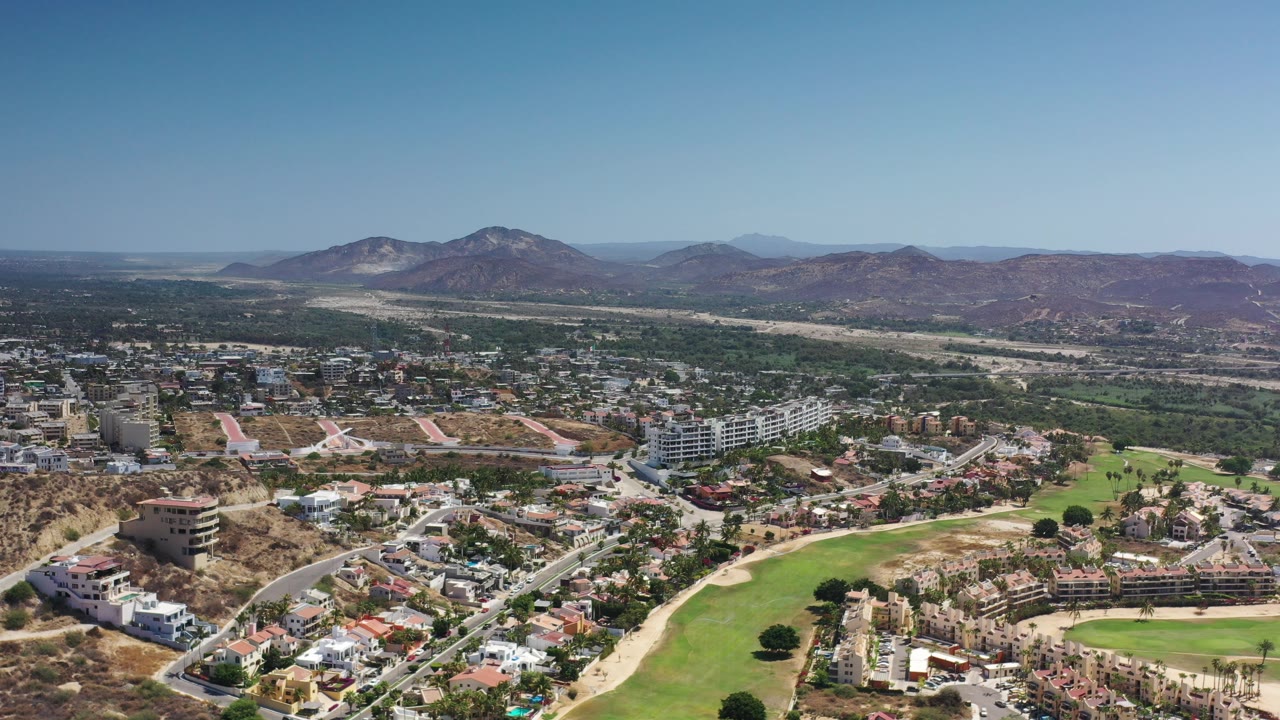 Wide aerial over residential streets, golf-course green, buildings, and mountains.