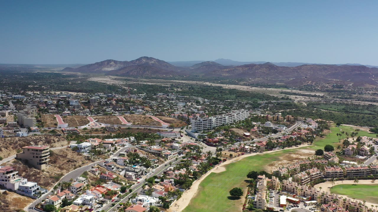 Aerial view of roads, buildings, green fields, and mountains under blue sky.