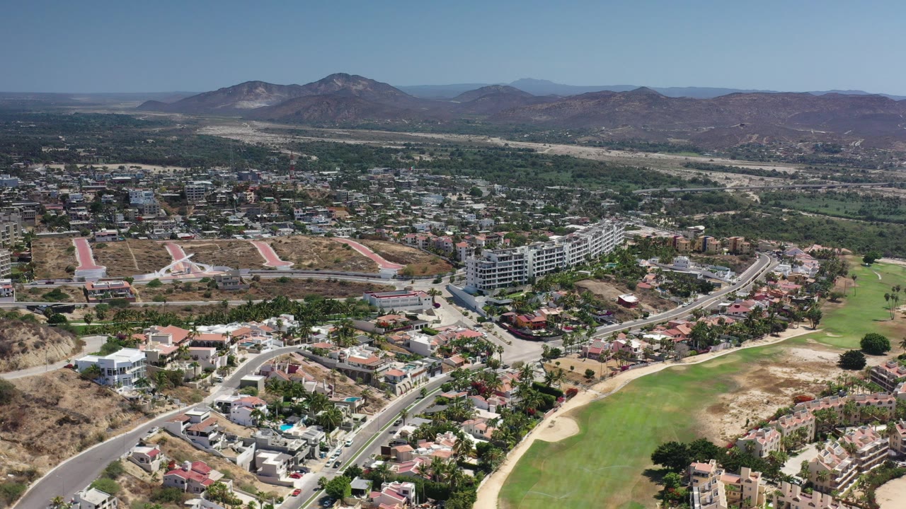 Opening aerial frame over Los Cabos showing residential streets, green fields, mid-rise buildings, dry hills, distant mountains, and no visible people.
