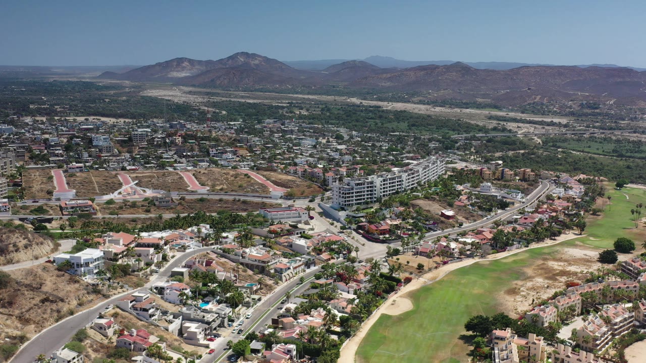 Aerial frame in Los Cabos with curved roads and homes in the foreground, green fairway-like fields in the middle distance, mountains on the horizon, and no visible people.