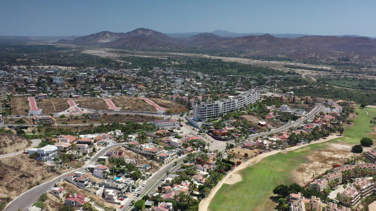Drone-height view of buildings, green fields, scrubland, and distant mountains.