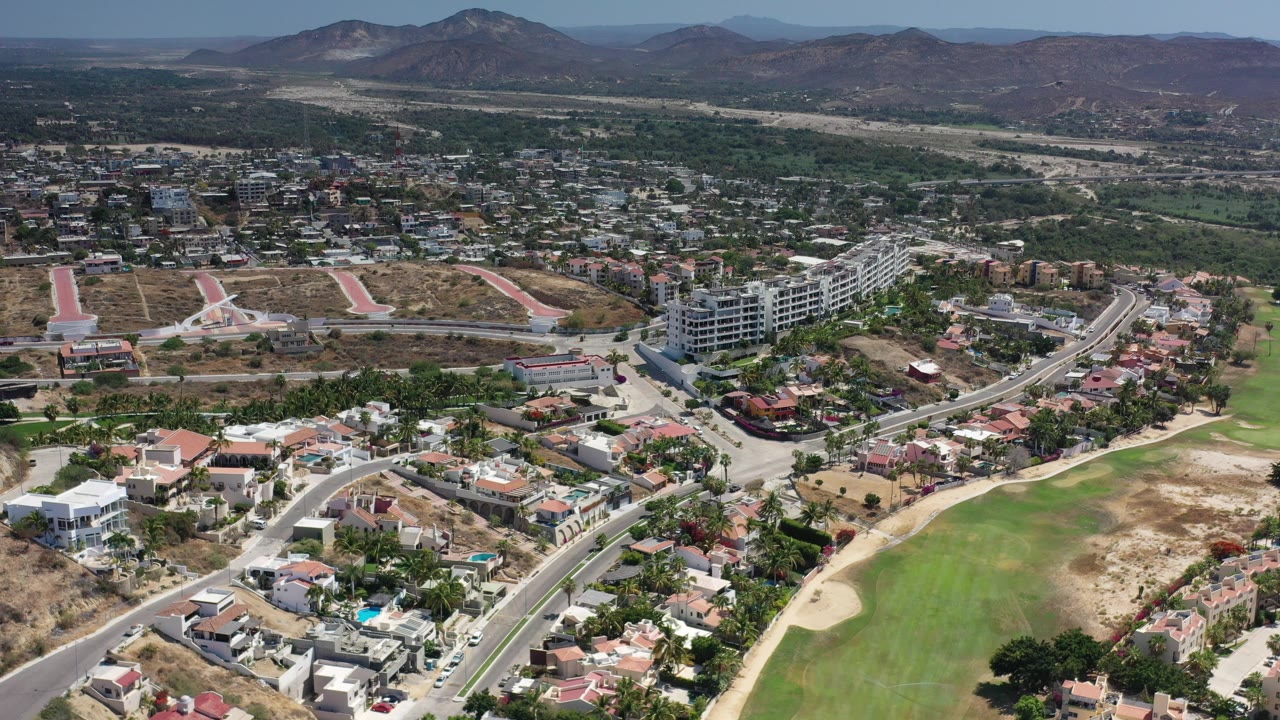 Opening aerial frame over Los Cabos, Mexico, with residential homes, a curving road, a green fairway-like strip, dry lots, distant town, and no recognizable people.