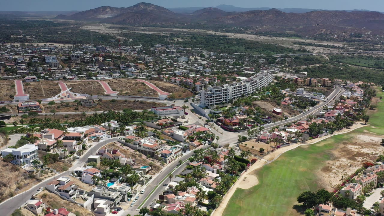 Aerial view in Los Cabos, Mexico, looking across houses, a pale road network, a broad green landscaped area, dry ground, distant buildings, and low mountains.