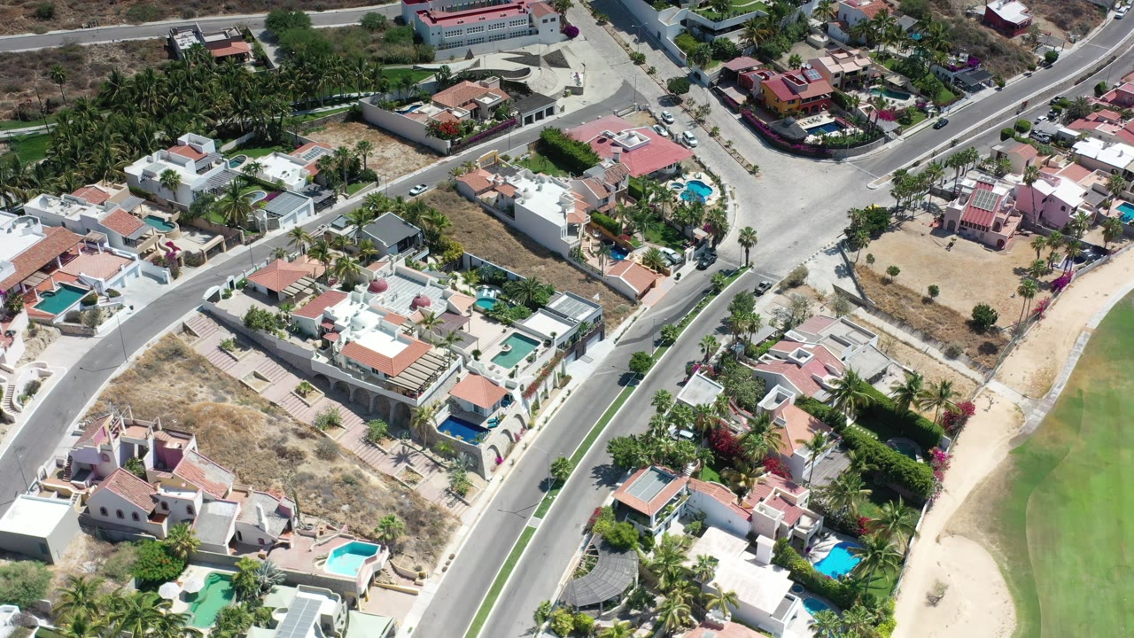 Aerial Los Cabos frame showing white and terracotta-roofed homes, blue pools, palms, a main road, dry lots, and no visible people.