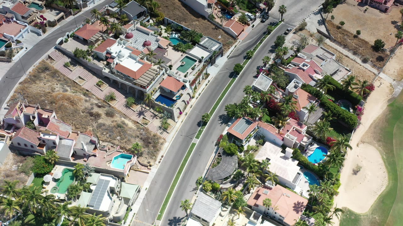 Opening aerial frame over Los Cabos, Mexico, showing a divided residential road, walled homes, turquoise pools, palm trees, dry lots, green fairway, sand bunkers, and no visible people.