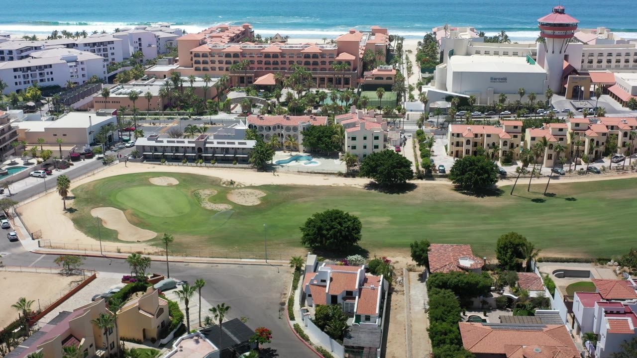 Los Cabos aerial view with putting greens and sand traps below, palm-lined streets and resort buildings ahead, ocean surf at the horizon, and no visible people.