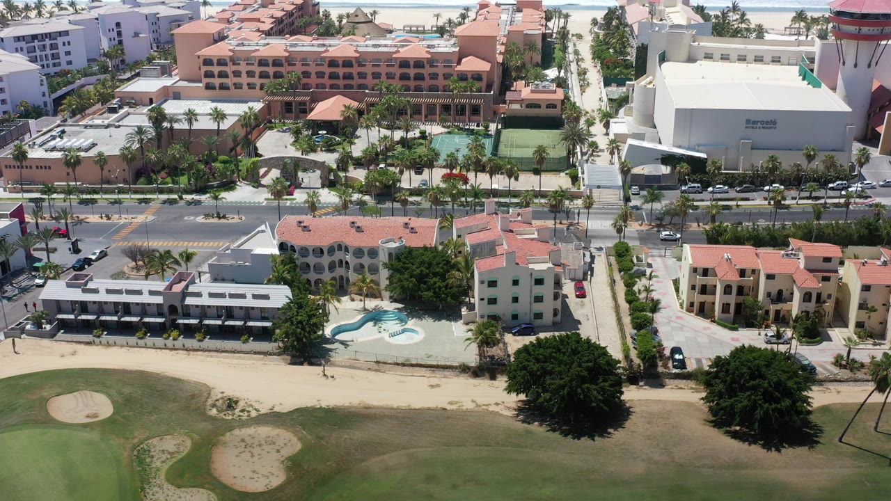 Opening aerial frame over Los Cabos resort buildings with red roofs, blue pools, a palm-lined road, and a dry golf course edge in the foreground, with no visible people.