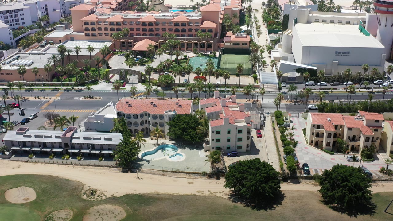 Overhead resort view with large white building, road, palm trees, pools, and golf course.