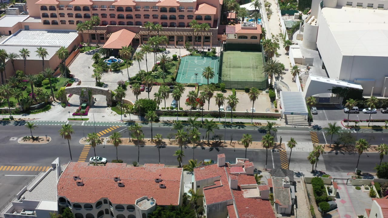 Aerial frame in Los Cabos, Mexico with a blue fountain, palm landscaping, green basketball court, fenced tennis court, large white building, and no visible people.