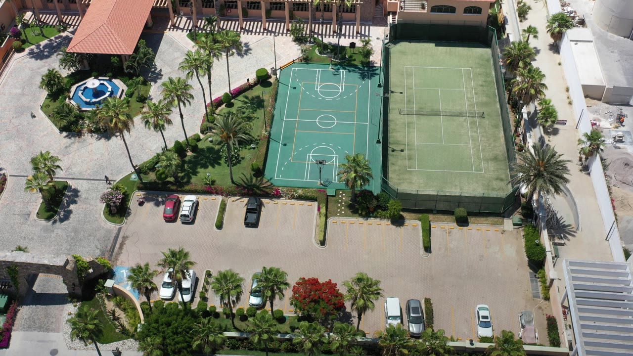Overhead aerial frame in Los Cabos, Mexico showing a green basketball court, tennis court, palm landscaping, parked cars, terracotta roofs, and no visible people.