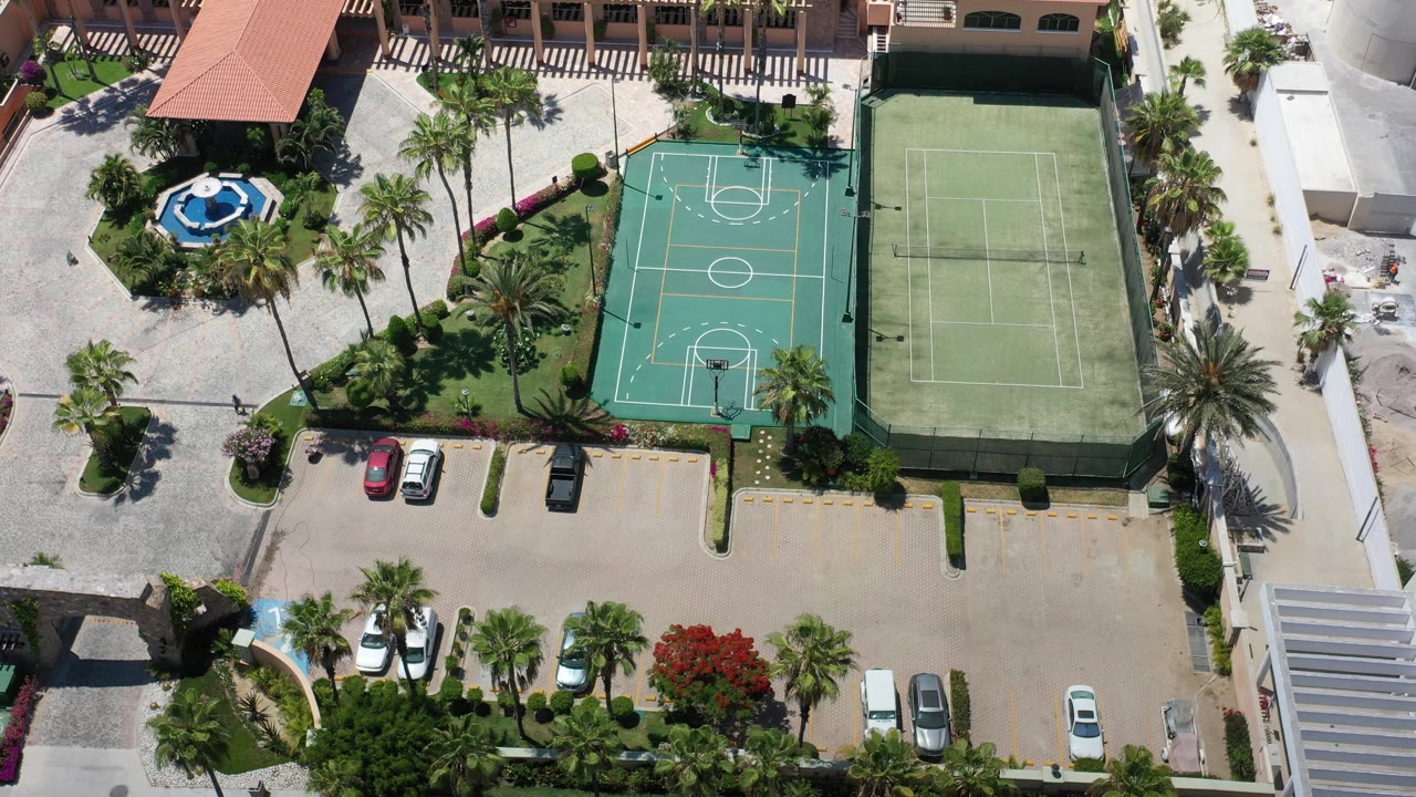 Straight-down Los Cabos aerial showing the basketball court and tennis court more centered, with palms, roofs, parked cars, paved areas, and no visible people.