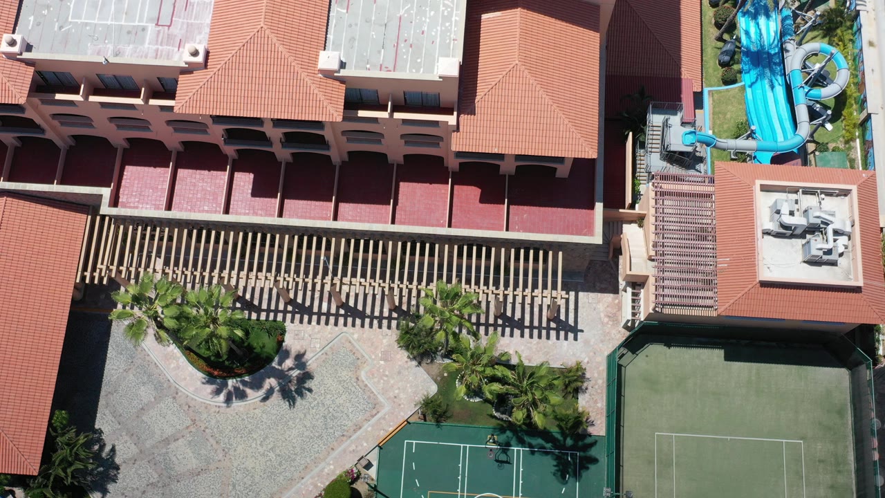 Top-down aerial frame over a Los Cabos resort showing red tile roofs, a small tennis court, palm trees, and turquoise water slides, with no visible people.