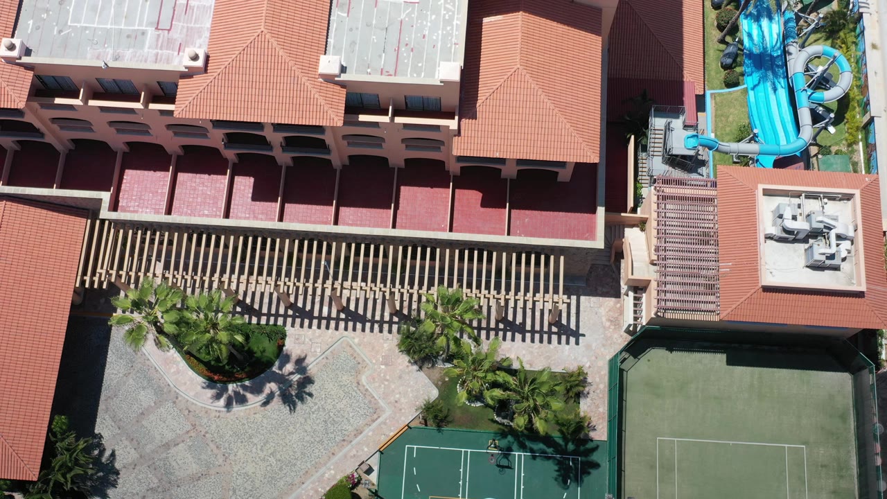 Aerial view in Los Cabos centered on a resort roofline and striped pergola shadows, with a tennis court edge, palms, and bright blue water slides visible, with no visible people.
