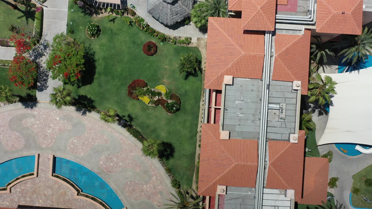 Top-down view in Los Cabos, Mexico showing red tile resort roofs, green lawns, curved paths, and partial blue pool areas with no visible people.