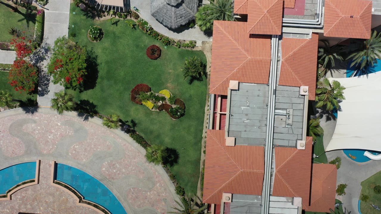Overhead Los Cabos resort frame with a long building roof centered between lawns, walkways, garden beds, and partial swimming pools with no visible people.