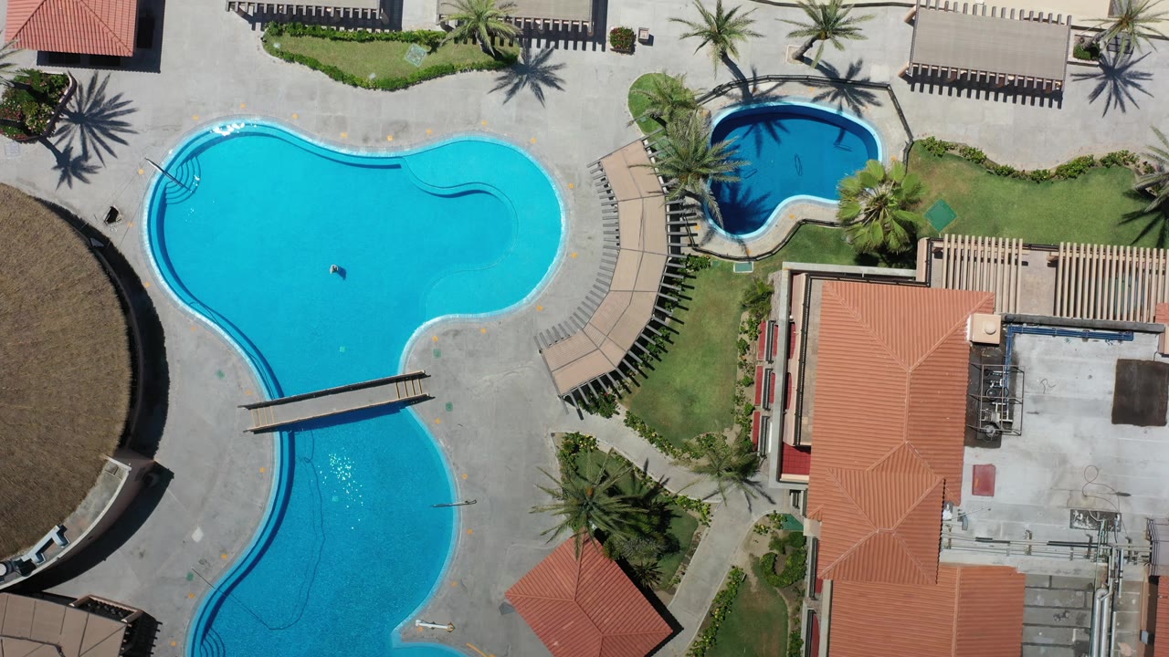 Overhead Los Cabos frame with the main blue swimming pool, a small adjacent pool, red roof structures, palm landscaping, lounge areas, and pale beach sand at the top right.