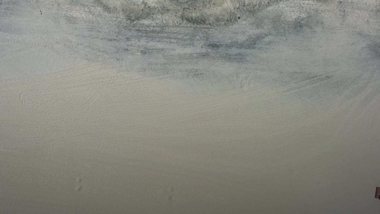 Top-down view of a Los Cabos beach with pale wet sand, muted foam traces near the top edge, and no visible people.