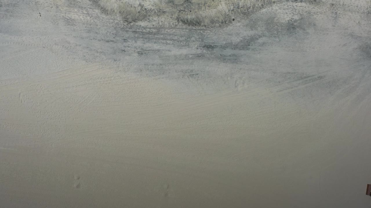 Overhead Los Cabos shoreline frame showing gray-white foam marks across tan sand, with no visible people or buildings.