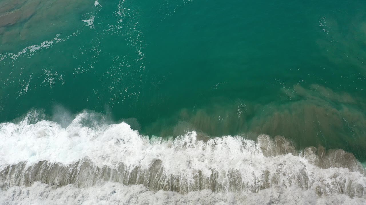 Overhead view in Los Cabos, Mexico of green ocean water above a foamy white surf line crossing wet sand, with no visible people.