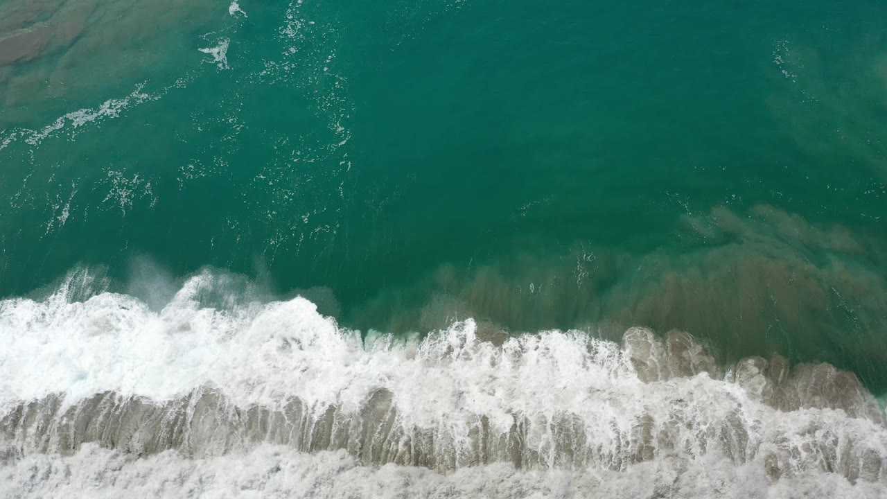Top-down Los Cabos, Mexico frame with white foam thickening across the center and green water filling the upper area, with no visible people.