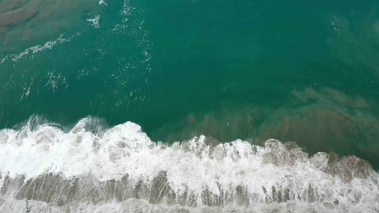 A larger white wave rolls through green coastal water from above.