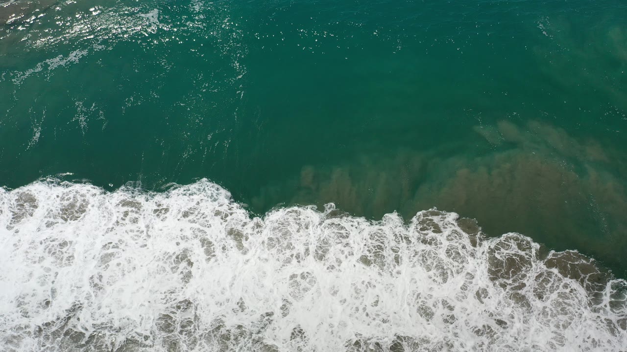 Overhead view of teal ocean and foamy surf over sandy water in Los Cabos, Mexico, with no visible people.
