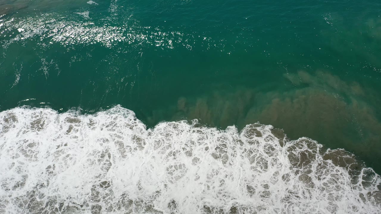 Aerial view of surf foam pushing across green-blue ocean water.